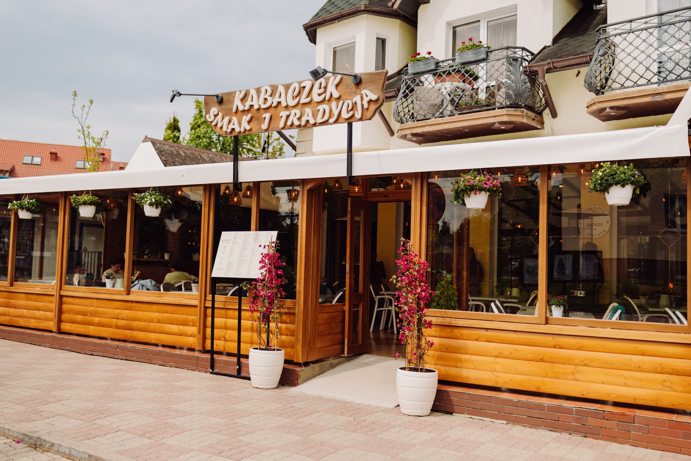 Restauracja Kabaczek w Ustroniu Morskim od frontu — drewniany szyld, ogródek z kwiatami i błękitne niebo